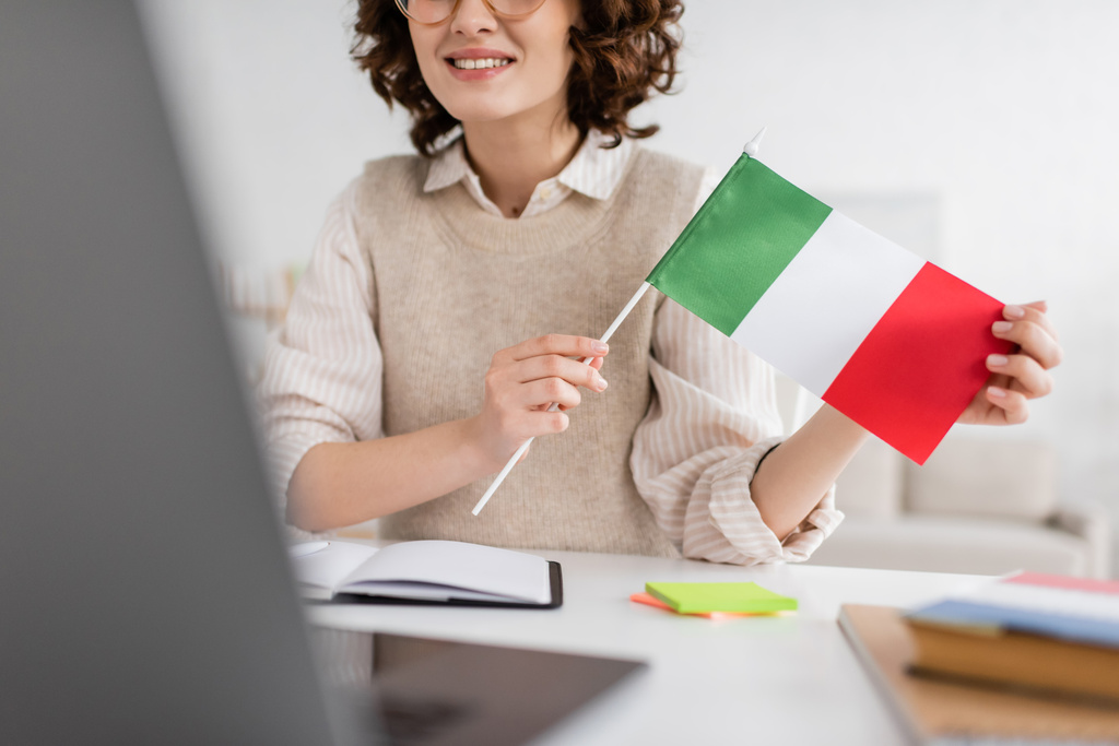 Evde bulanık dizüstü bilgisayarla ilgili çevrimiçi derste elinde İtalya bayrağı tutan gülümseyen dil öğretmeninin kısmi görüntüsü - Fotoğraf, Görsel