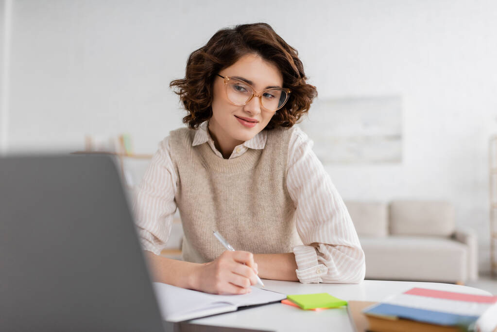 donna sorridente in occhiali che prende appunti mentre impara la lingua straniera e guarda lezione online sul computer portatile  - Foto, immagini