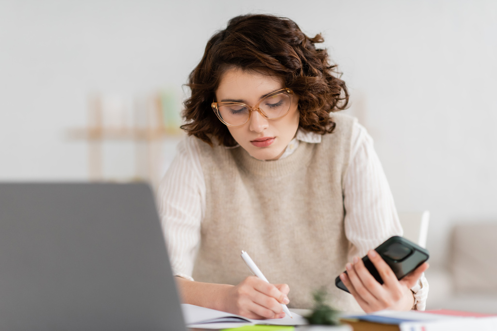 όμορφη φοιτήτρια σε γυαλιά κρατώντας smartphone, ενώ κρατώντας σημειώσεις κοντά θολή φορητό υπολογιστή στο γραφείο  - Φωτογραφία, εικόνα