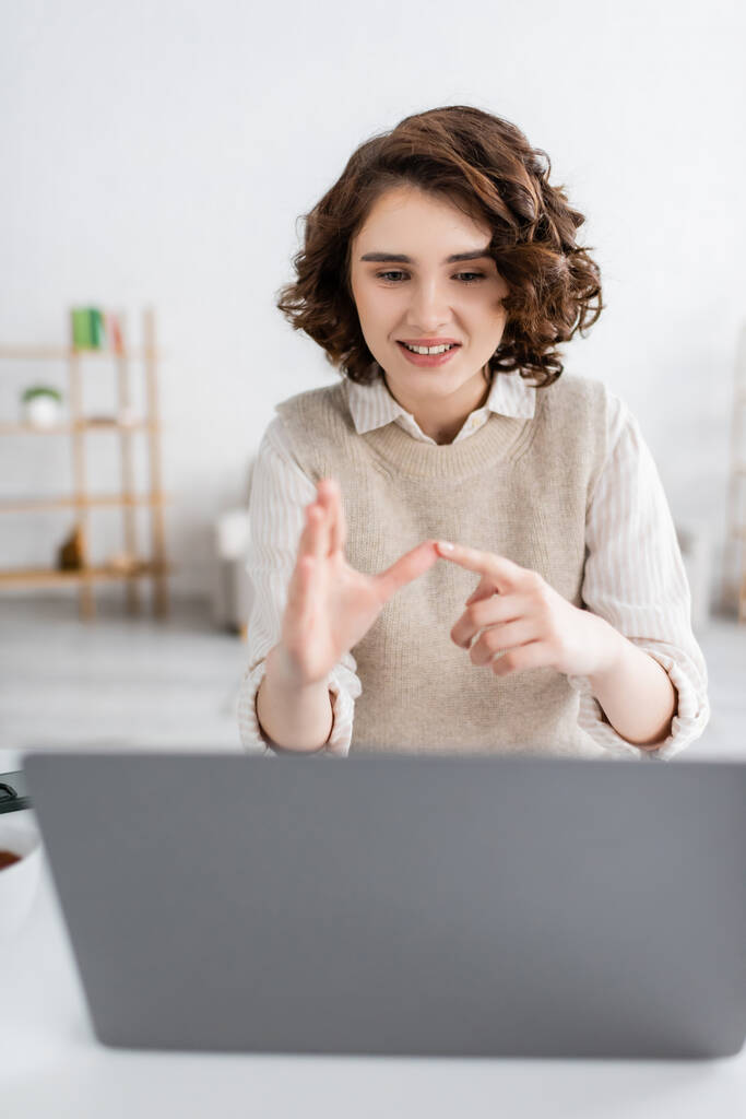 mulher alegre com cabelo encaracolado ensino língua de sinais alfabeto perto de laptop borrado em casa - Foto, Imagem