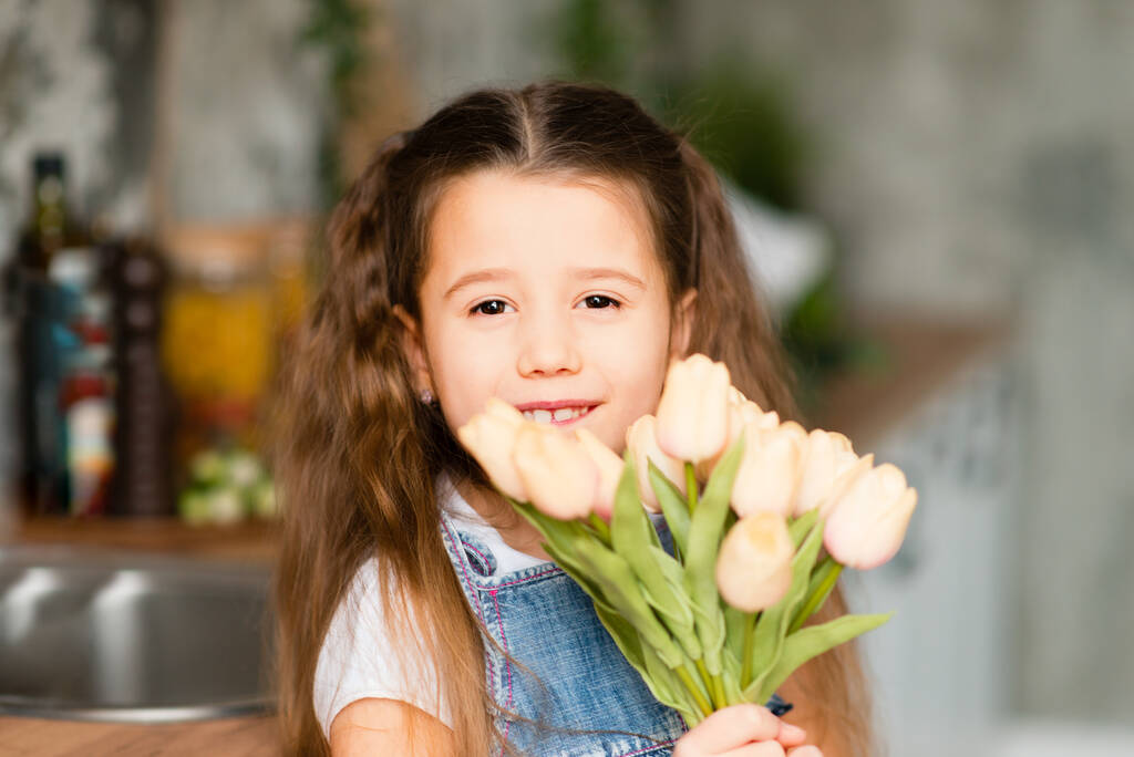 Smiling child gives flowers. portrait of a little girl with a bouquet of tulips flowers. girl in a denim dress. Gray interior. Mother's Day, birthday. - Photo, Image