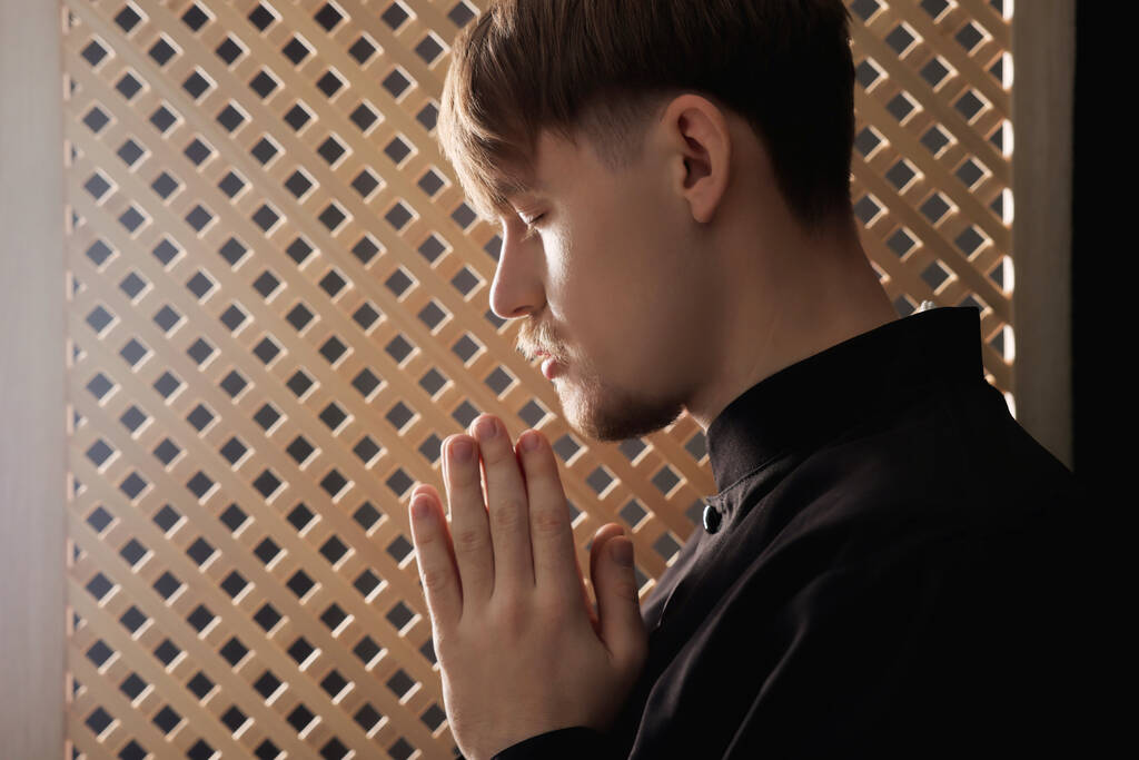 Καθολικός ιερέας προσεύχεται κοντά σε ξύλινο παράθυρο στο εξομολογητήριο περίπτερο - Φωτογραφία, εικόνα