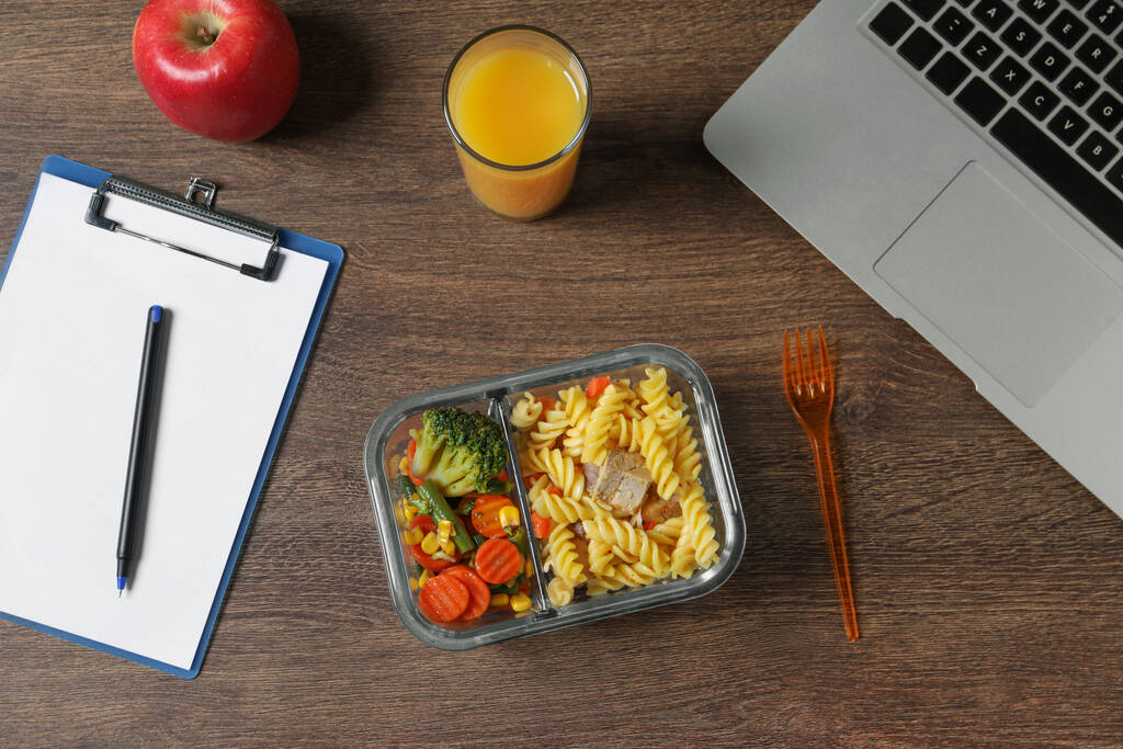 Behälter mit leckerem Essen, Gabel, Laptop, Saftglas, Apfel und Klemmbrett auf Holztisch, flach gelegt. Geschäftsessen - Foto, Bild