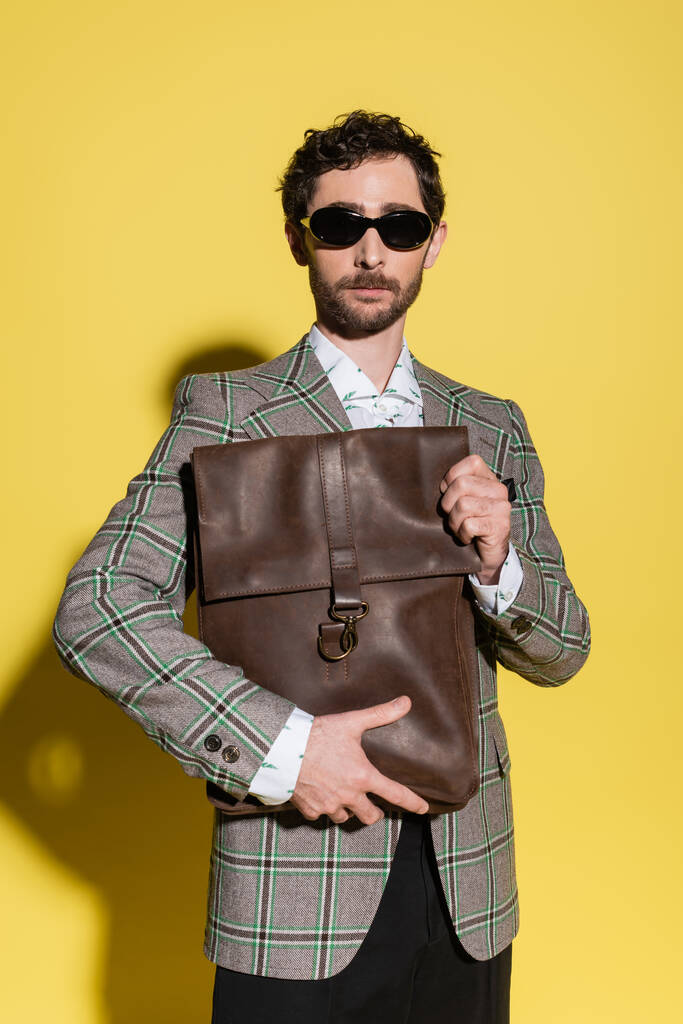 Charming guy in sunglasses and blazer holding bag on yellow background  - Photo, Image