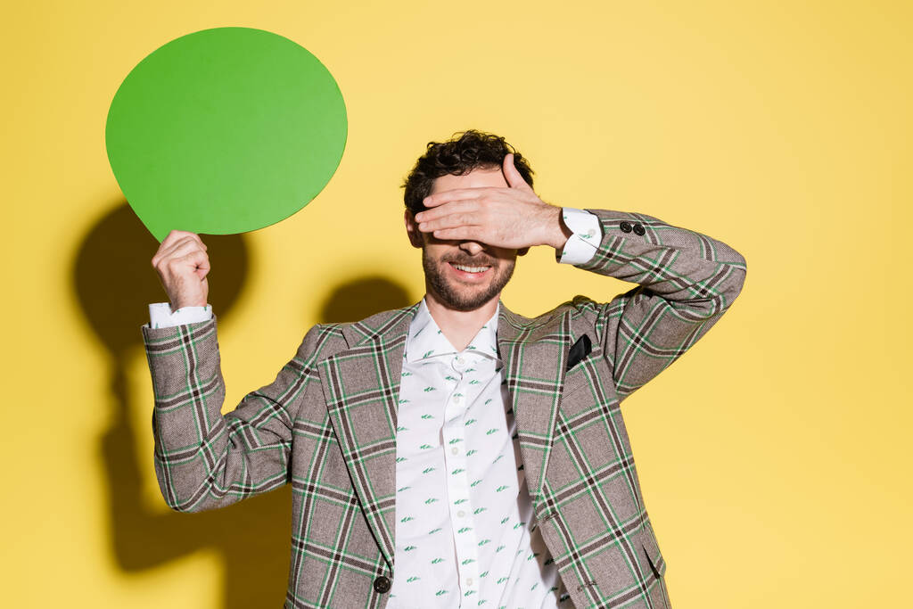 Smiling model covering eyes while holding speech bubble on yellow background  - Photo, Image