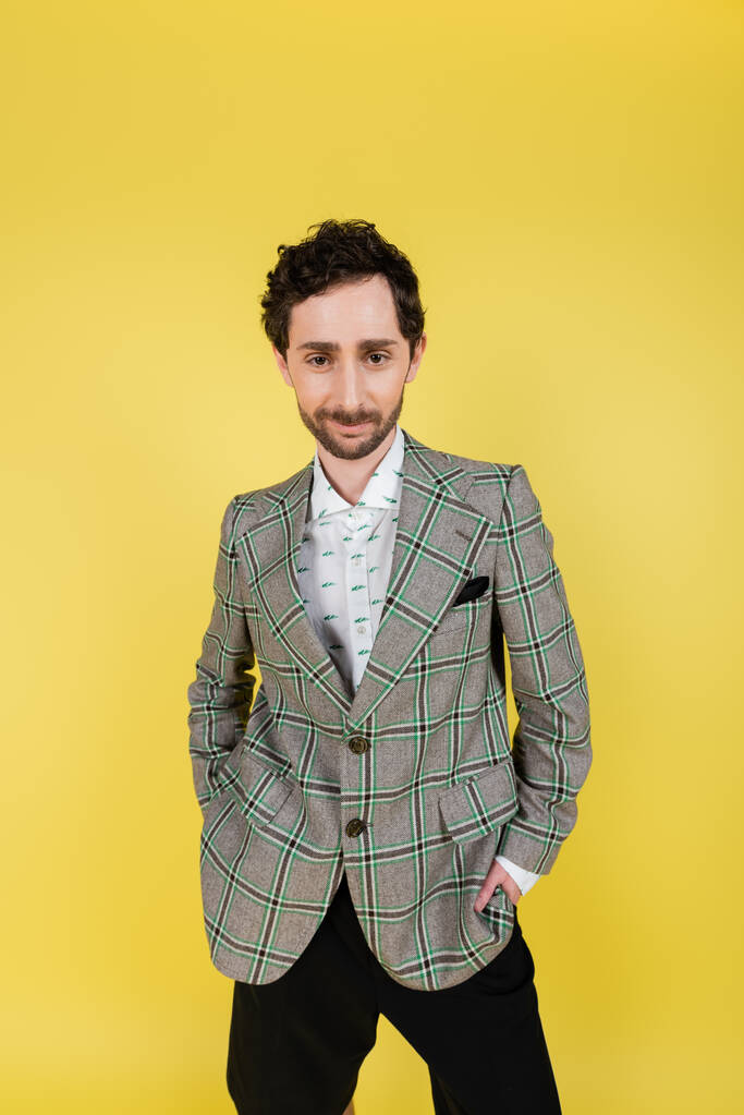Stylish man in shirt and checkered jacket posing with hands in pockets isolated on yellow   - Photo, Image