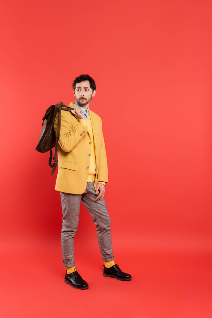 Longitud completa del modelo elegante en blazer amarillo que sostiene la mochila sobre fondo rojo coral - Foto, Imagen