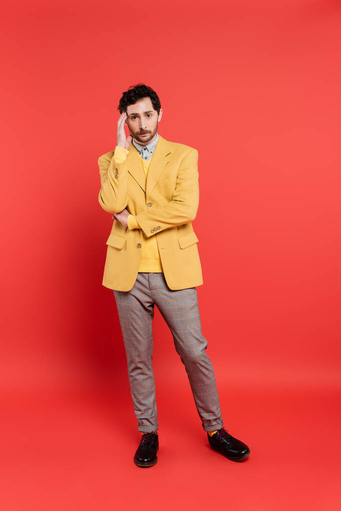 volledige lengte van gestresste man in gele blazer en grijze broek raken voorhoofd terwijl staan op rood koraal achtergrond - Foto, afbeelding