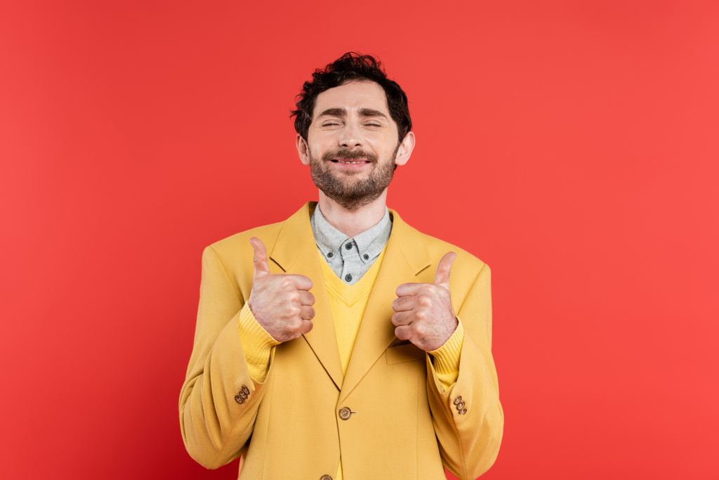 pleased man in yellow blazer standing with closed eyes and showing thumbs up on red coral background - Photo, Image