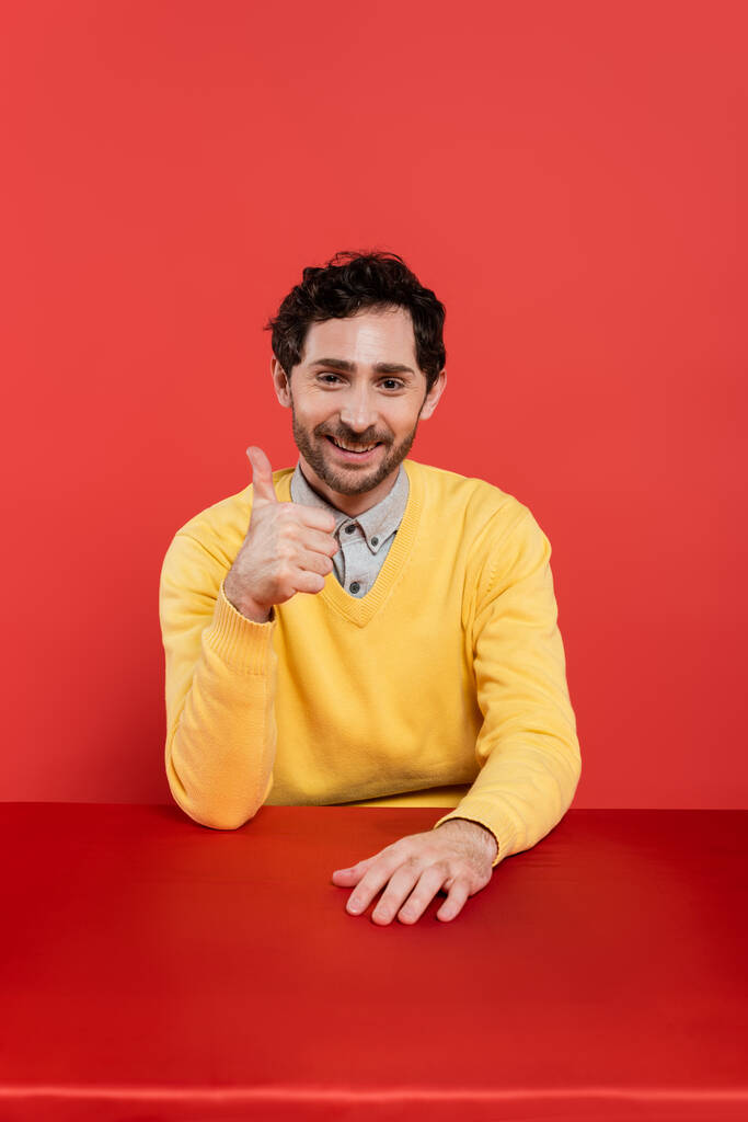 szczęśliwy człowiek w żółtym swetrze z długim rękawem pokazuje kciuk w górę na czerwonym tle korala  - Zdjęcie, obraz