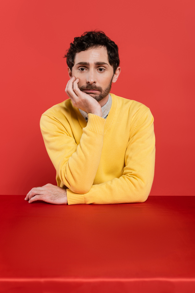 uomo frustrato in giallo maglione a maniche lunghe seduto alla scrivania su sfondo corallo rosso  - Foto, immagini