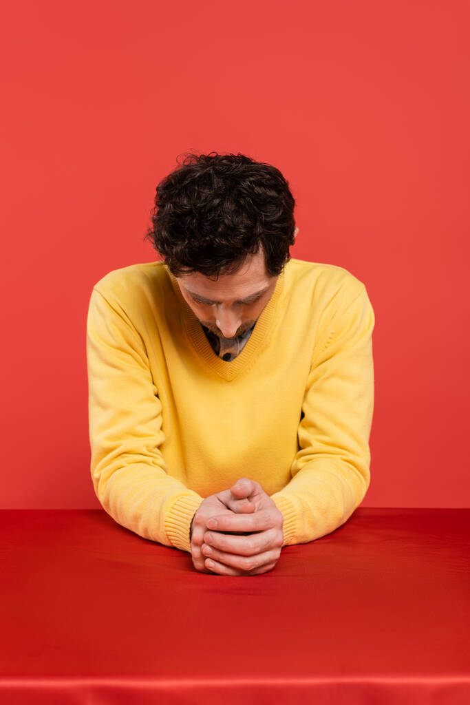 man in yellow long sleeve jumper sitting with bowed head at table isolated on coral background  - Photo, Image