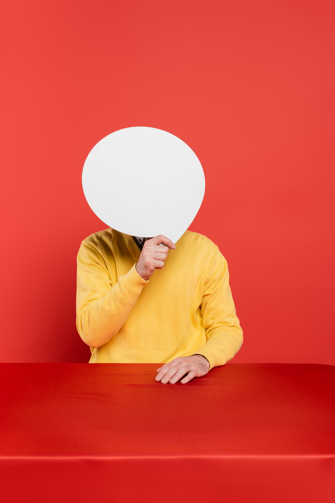 man in yellow long sleeve jumper covering face with blank speech bubble on red coral background  - Photo, Image
