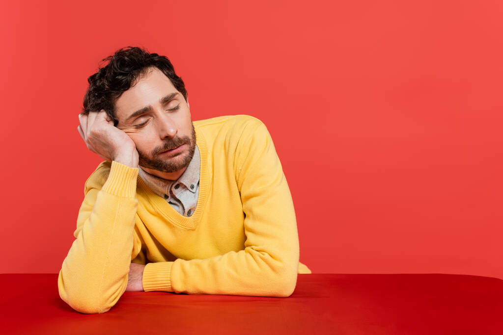 zmęczony mężczyzna w żółtym swetrze z długim rękawem śpi przy biurku odizolowany na koralowym tle  - Zdjęcie, obraz