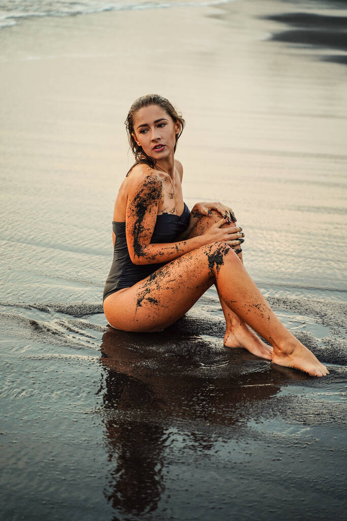 Sexy Frau im Badeanzug sitzt am schwarzen Strand. Gebräunte Frau mit perfekter Körperform. Entspannen und natürliche Körperbehandlung. - Foto, Bild