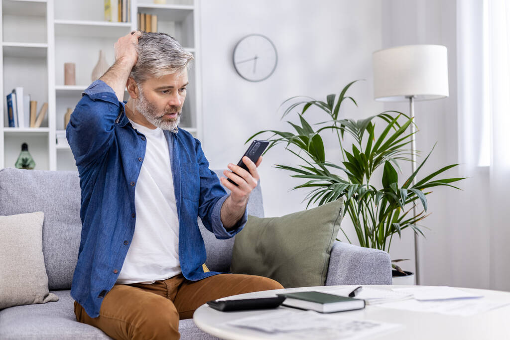Взрослый мужчина, сидящий дома один на диване, пользующийся смартфоном в гостиной, читающий плохие новости онлайн из приложения по телефону, расстроенный и нервный. - Фото, изображение