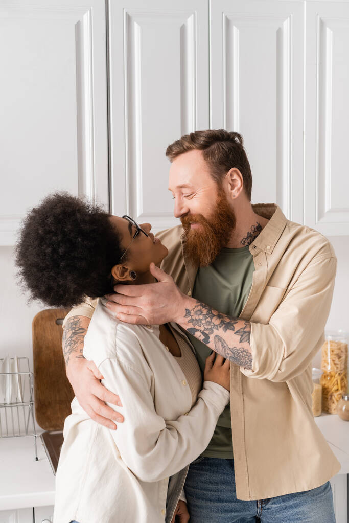Positive tattooed man touching african american girlfriend in kitchen at home  - Photo, Image