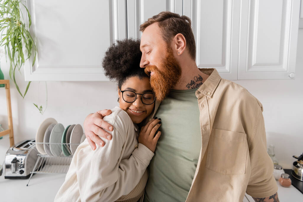 Ενθουσιασμένος άντρας αγκαλιάζει την Αφροαμερικανή κοπέλα του με γυαλιά στην κουζίνα  - Φωτογραφία, εικόνα