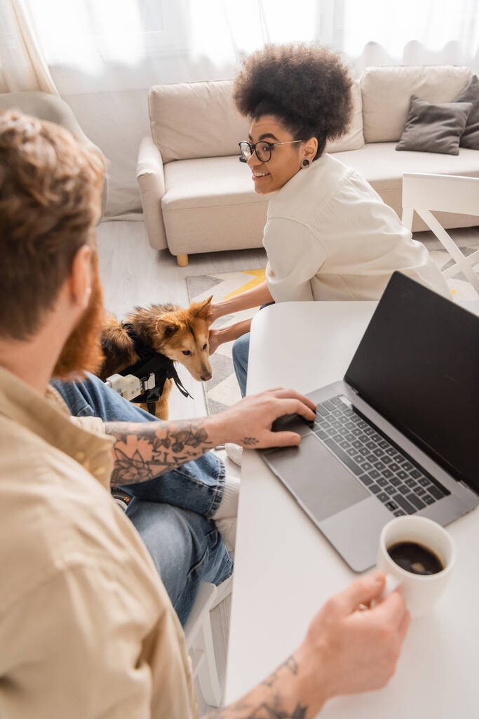 Cheerful african american woman petting handicapped dog near blurred boyfriend with laptop and coffee at home  - Photo, Image