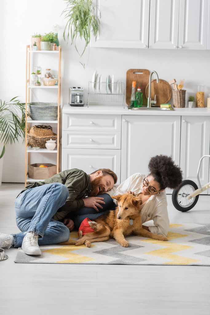 Šťastný mezirasový pár smějící se a sedí v blízkosti zdravotně postiženého psa na podlaze doma  - Fotografie, Obrázek