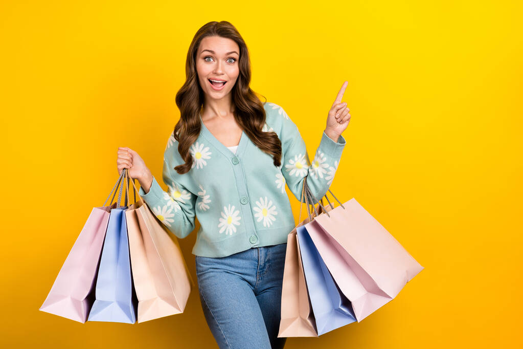 Foto van jonge verbaasd dame dragen stijlvolle bloemen print vest directe vinger lege ruimte shopping tassen verkoop geïsoleerd op gele kleur achtergrond. - Foto, afbeelding