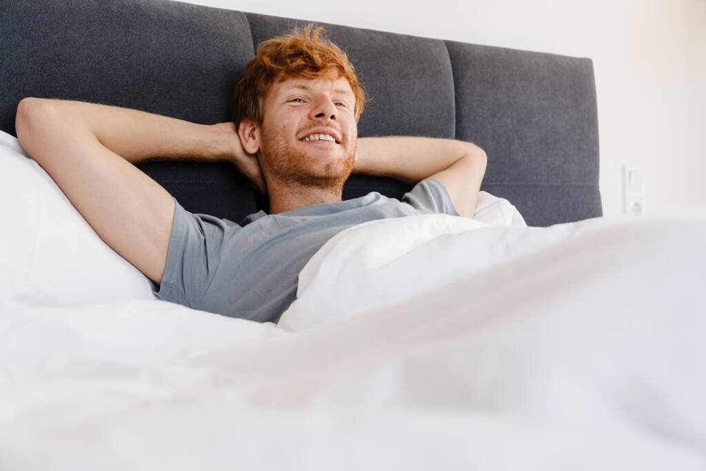 Νεαρός όμορφος χαμογελαστός κοκκινομάλλης με γκρι μπλουζάκι με τα χέρια πίσω από το κεφάλι του κοιτάζοντας στην άκρη ξαπλωμένος στο κρεβάτι στο σπίτι - Φωτογραφία, εικόνα