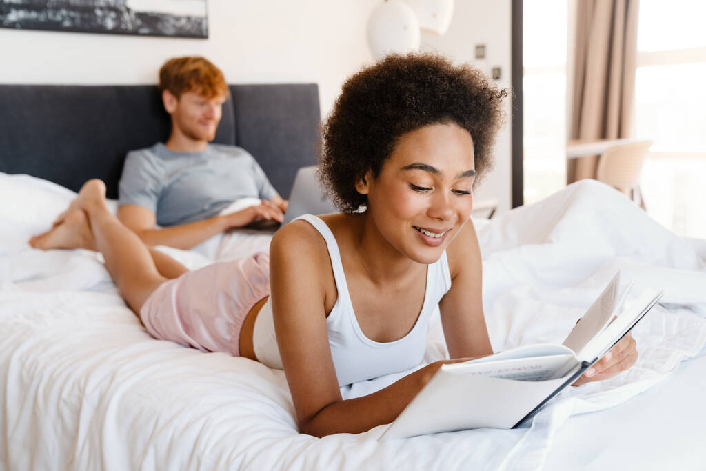 Młoda, piękna afrykańska, kręcona, uśmiechnięta kobieta czyta książkę ze swoim chłopakiem, który pracuje z laptopem w łóżku w domu - Zdjęcie, obraz