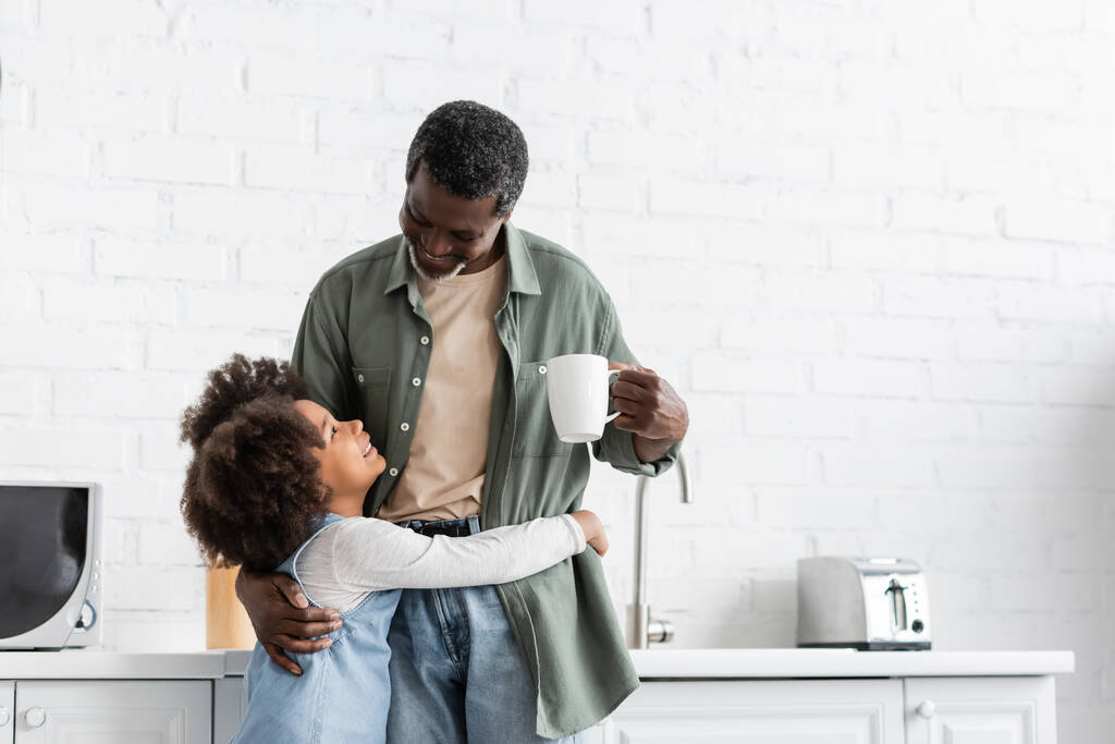 boldog afro-amerikai gyerek ölelés nagyapa kezében csésze kávé a konyhában  - Fotó, kép