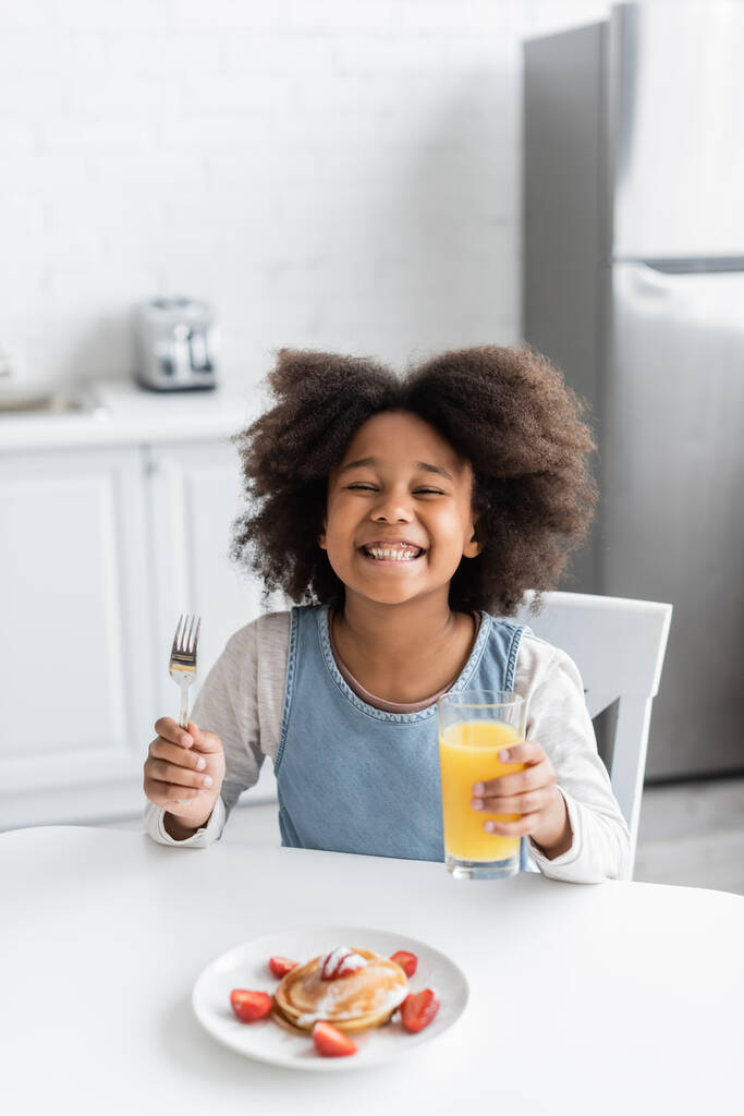 chica afroamericana feliz sosteniendo tenedor y vaso de jugo de naranja cerca de panqueques mientras disfruta del desayuno  - Foto, Imagen