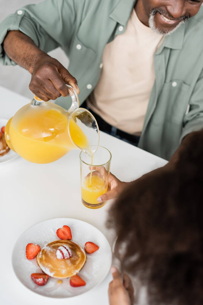 top view of happy african american man pouring orange juice near curly granddaughter during breakfast  - Photo, Image