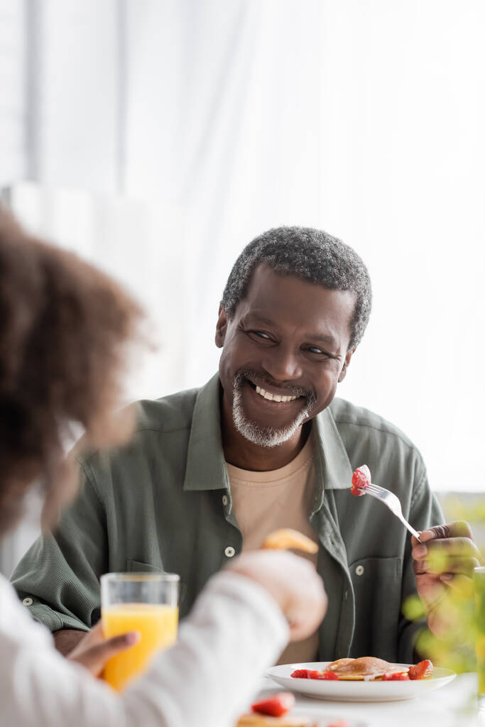 Mutlu Afrikalı Amerikalı adam elinde çatalla taze çilek tutuyor ve kahvaltıda torununa bakıyor.  - Fotoğraf, Görsel