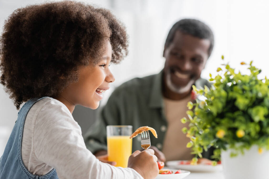 feliz niño afroamericano feliz sosteniendo tenedor con panqueque mientras desayuna con el abuelo  - Foto, imagen