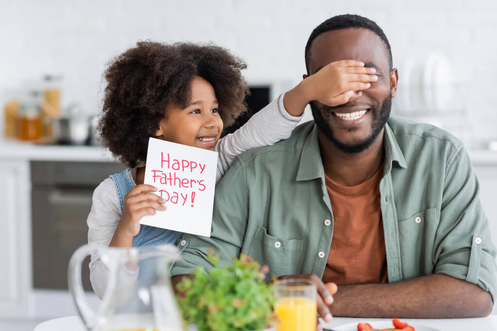 Kıvırcık Afrikalı Amerikalı kız elinde mutlu babalar günü kartı tutarken babasının gözlerini kapatıyor.  - Fotoğraf, Görsel
