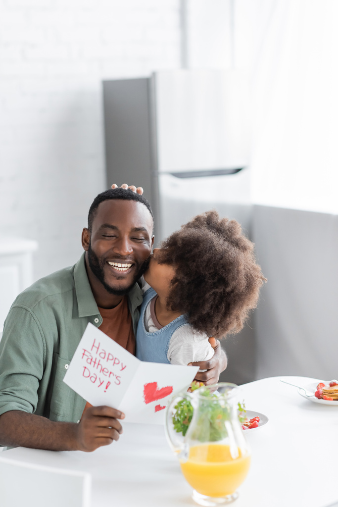 curly african american kid kissing cheek of happy father with greeting card on fathers day  - Photo, Image