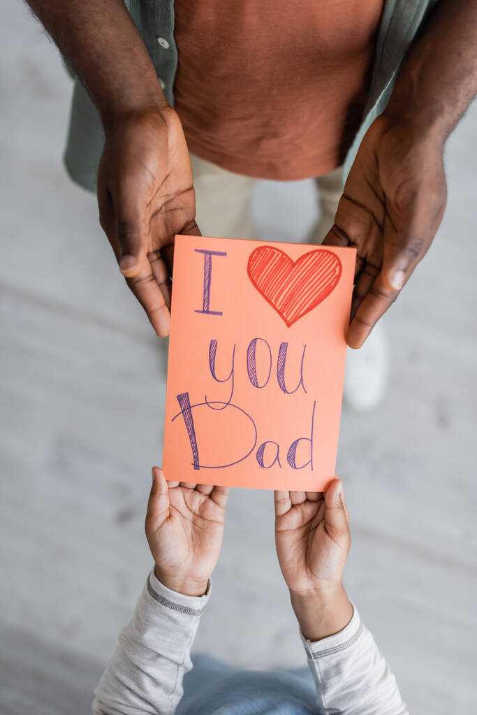 vista superior del niño afroamericano recortado sosteniendo la tarjeta de felicitación con te amo papá letras cerca del hombre en el día de los padres  - Foto, Imagen
