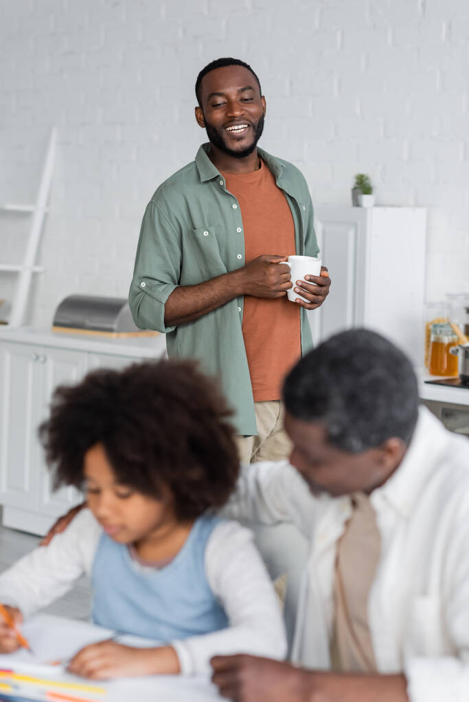 feliz africano americano hombre celebración taza y mirando hija dibujo cerca abuelo en borrosa primer plano  - Foto, imagen