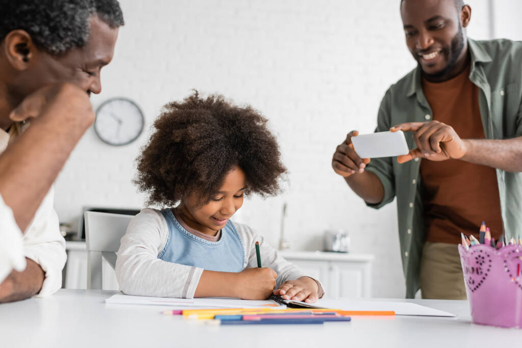 glücklicher afrikanisch-amerikanischer Mann beim Fotografieren auf dem Smartphone, während seine Tochter und sein Vater zusammen zeichnen  - Foto, Bild