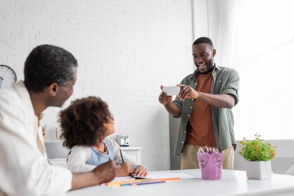 Neşeli Afrikalı Amerikalı adam, kızı ve babası birlikte resim çizerken akıllı telefondan fotoğraf çekiyor.  - Fotoğraf, Görsel
