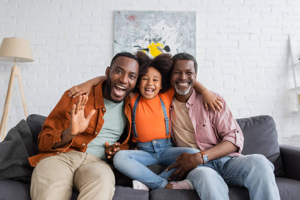 Весела афроамериканська дівчина, яка обіймає дідуся й тата на дивані у вітальні.  - Фото, зображення