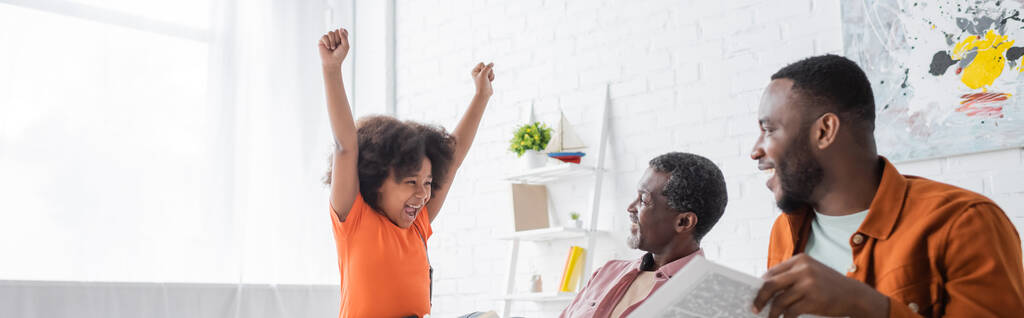 Fille afro-américaine excitée montrant oui geste près des parents avec journal à la maison, bannière  - Photo, image