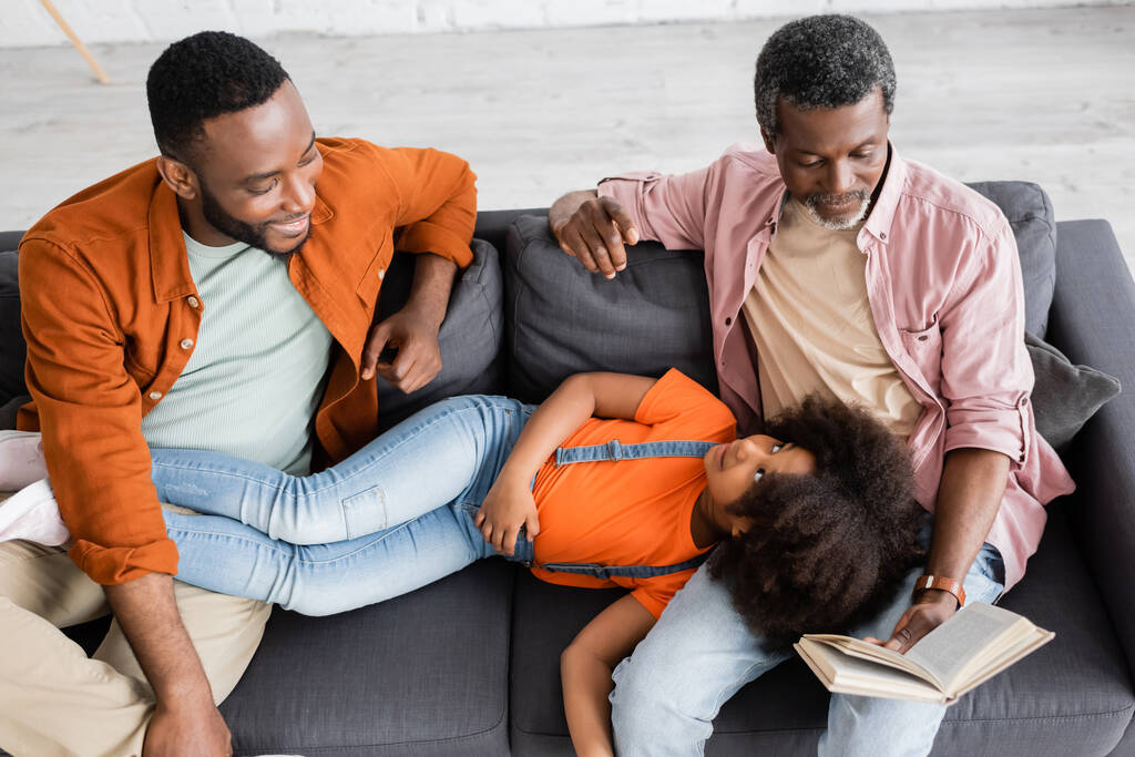 Висококутний вид на афро-американську дівчинку, що лежить поруч з батьком і дідусем з книжкою вдома.  - Фото, зображення