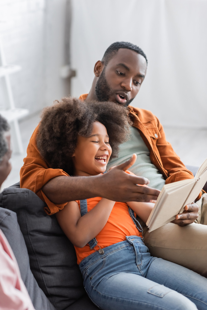 Afrykański Amerykanin czyta książkę w pobliżu wesołej córki i ojca w domu  - Zdjęcie, obraz