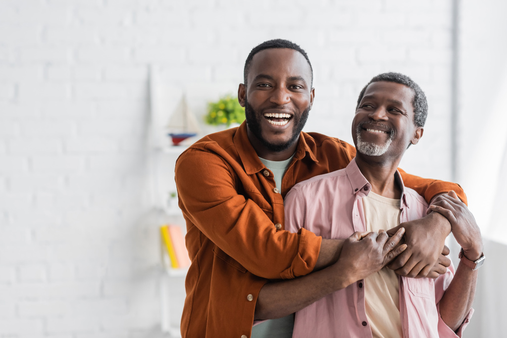 Vrolijke Afrikaanse Amerikaanse man knuffelen volwassen vader en kijken naar camera thuis  - Foto, afbeelding
