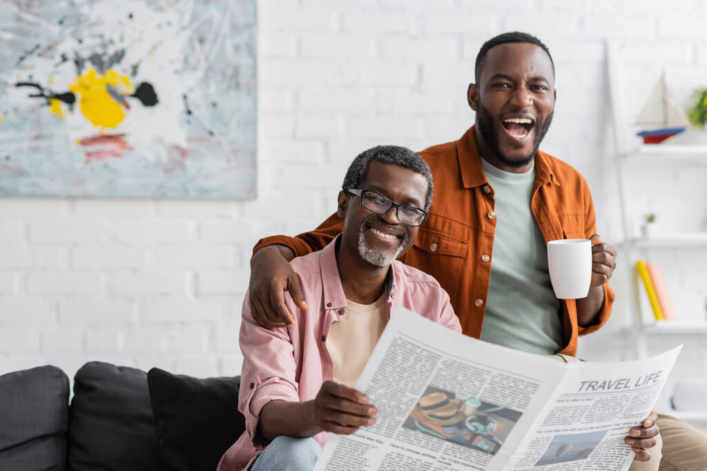 Beztroski Afrykanin amerykański człowiek z filiżanki przytulanie dojrzały ojciec z gazety w salonie  - Zdjęcie, obraz