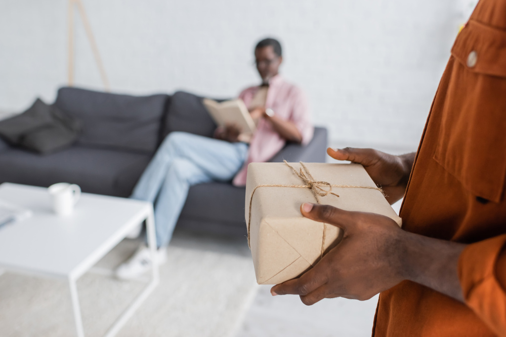 Uomo afroamericano con in mano una scatola regalo durante la festa del padre vicino al padre sfocato a casa - Foto, immagini