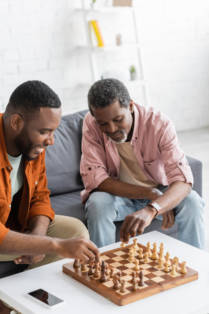 Uomo afroamericano che gioca a scacchi con il figlio sorridente vicino smartphone a casa  - Foto, immagini