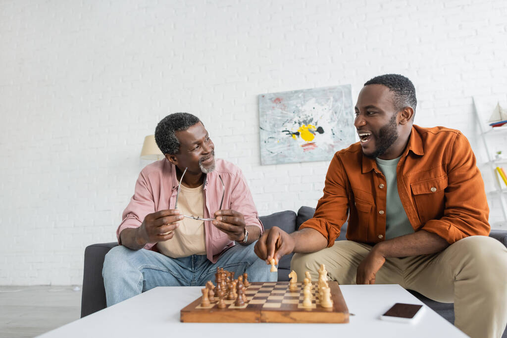 Overjoyed uomo africano americano giocare a scacchi con papà maturo vicino smartphone con schermo bianco a casa  - Foto, immagini