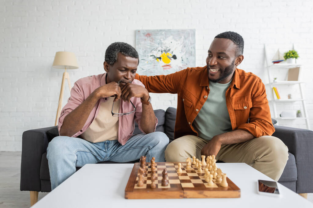 Smiling african american man hugging focused dad while playing chess in living room  - Photo, Image