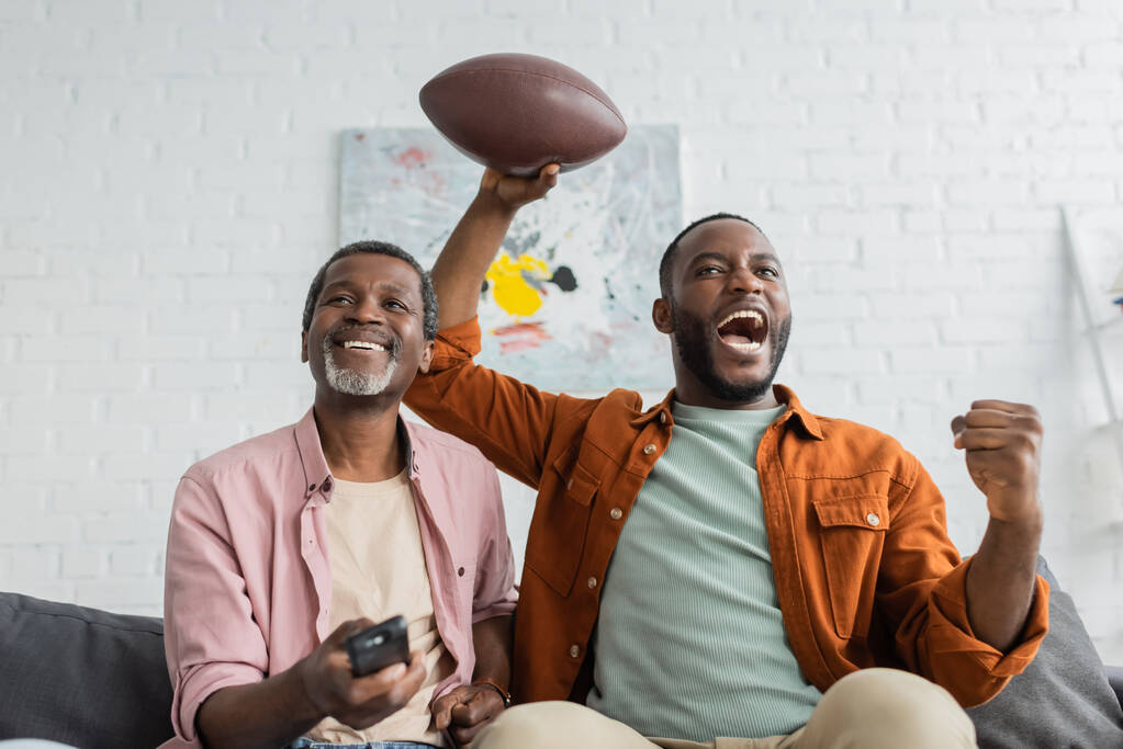 Emocionado afro-americano segurando bola de rugby perto do pai com controle remoto na sala de estar  - Foto, Imagem
