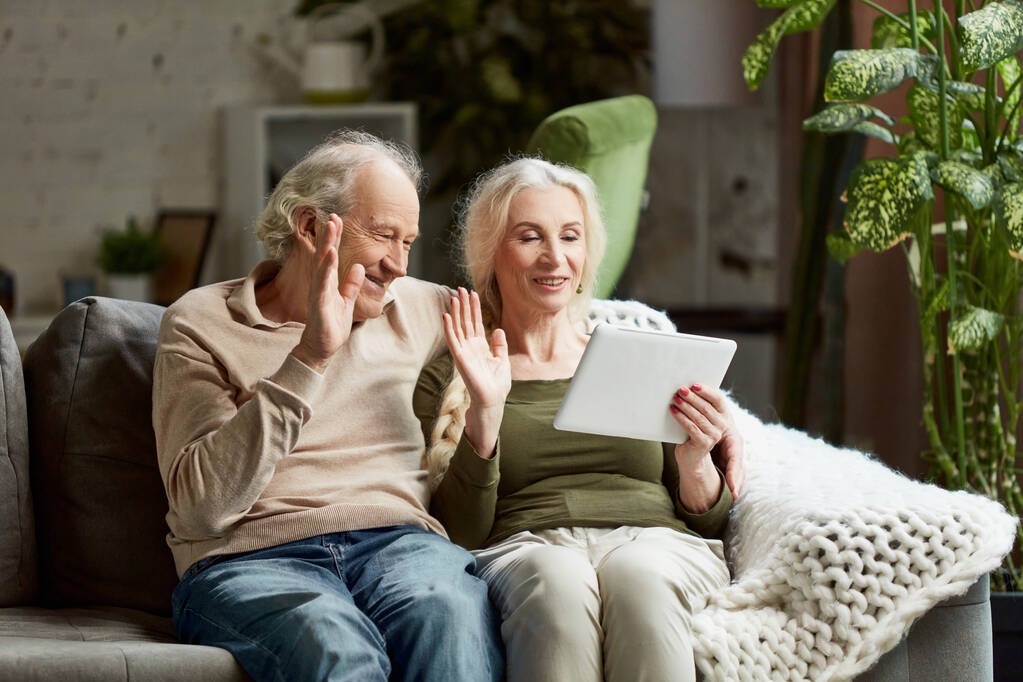 Avós, casal de idosos, homem e mulher sentados no sofá em casa e conversando em videochamada online com parentes. Usando tablet. Conceito de família, relacionamento, tecnologias modernas, comunicação - Foto, Imagem